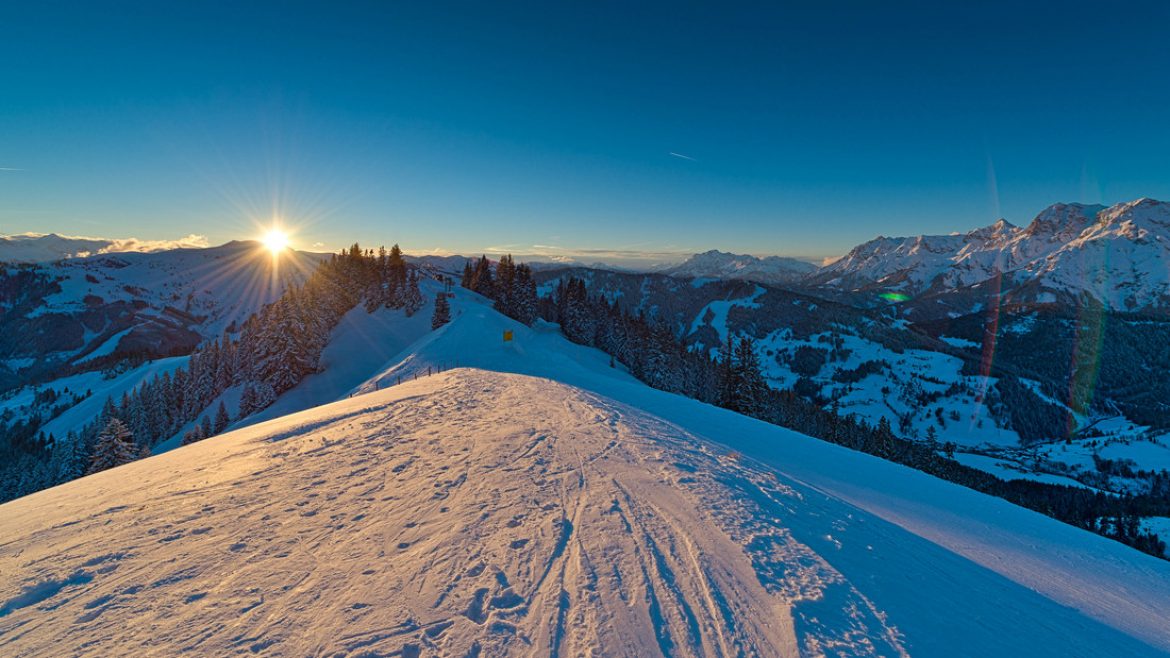Die Sonne geht über einem verschneiten Bergrücken unter und wirft lange Schatten auf den Schnee mit fernen Gipfeln im Hintergrund.
