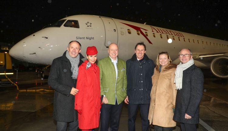 Sechs Personen posieren lächelnd vor einem Flugzeug der Austrian Airlines, das nachts auf einer nassen Landebahn steht.