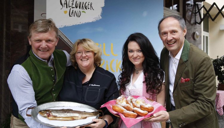 Vier lächelnde Menschen mit Tellern voller Fisch und Gebäck vor einem Salzburger Tourismusbanner.