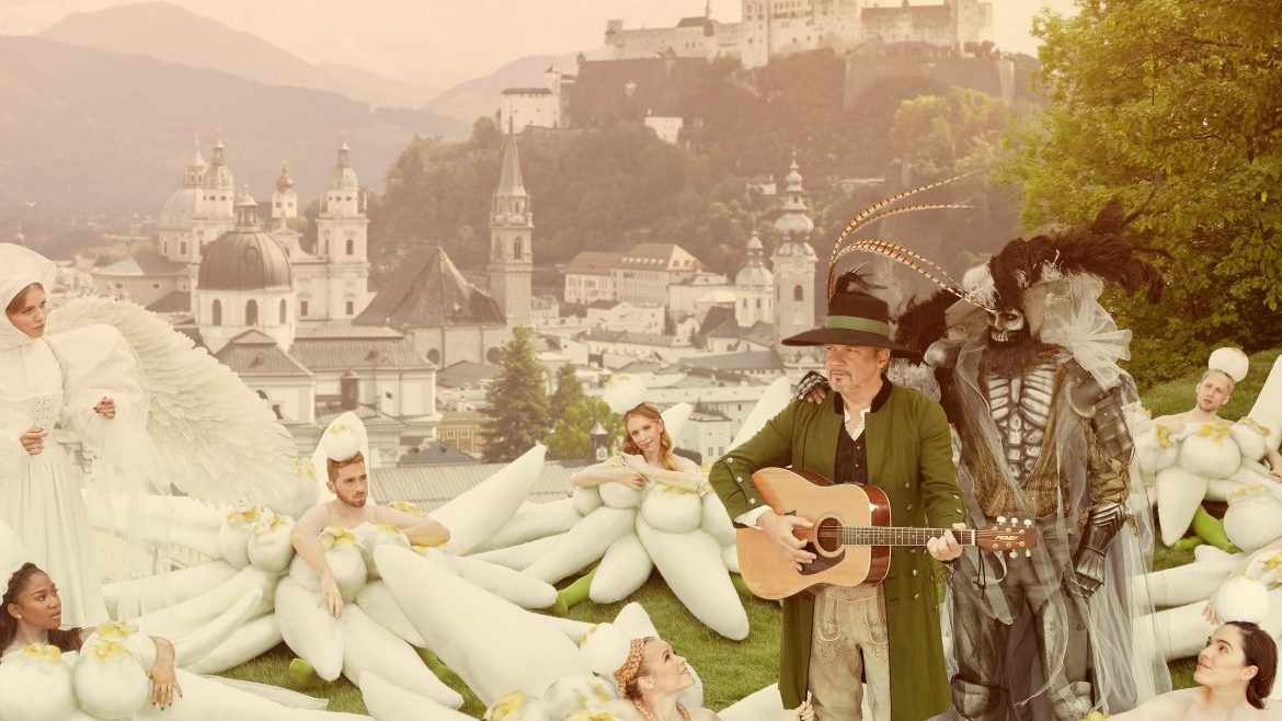 Menschen in skurrilen Kostümen, einige mit Federn, versammeln sich auf einem grasbewachsenen Hügel mit Blick auf eine Stadt und die Berge.