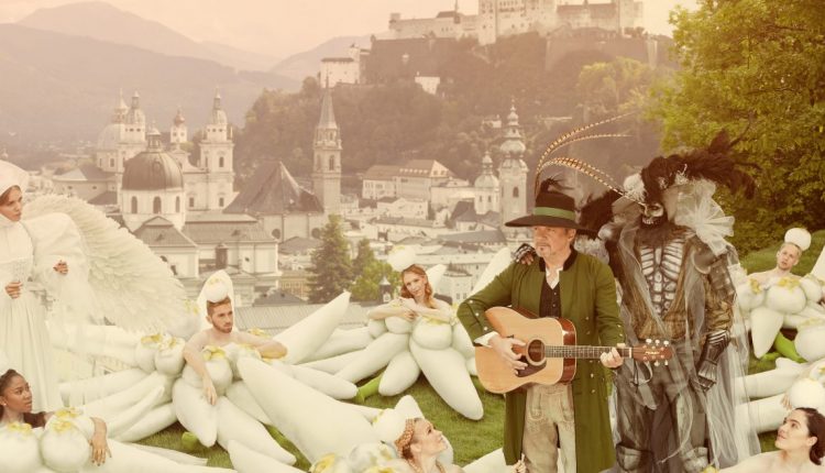 Menschen in skurrilen Kostümen, einige mit Federn, versammeln sich auf einem grasbewachsenen Hügel mit Blick auf eine Stadt und die Berge.