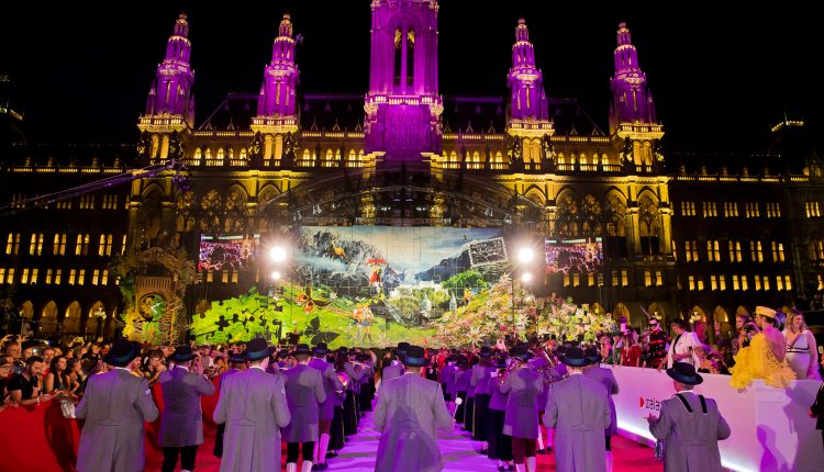 Eine Gruppe tritt in traditioneller Kleidung auf einem roten Teppich vor einem beleuchteten, prächtigen Gebäude bei Nacht auf.