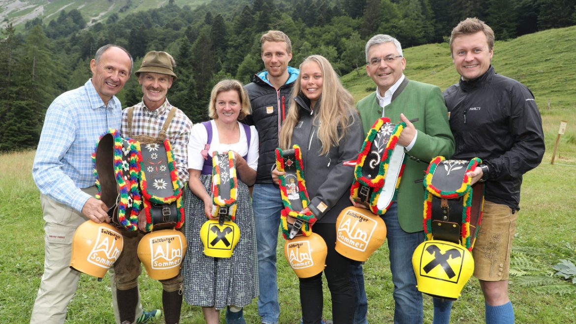 Sieben lächelnde Menschen im Freien, die große geschmückte Kuhglocken halten und legere Kleidung tragen, mit grünen Hügeln im Hintergrund.