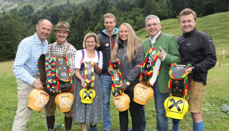Sieben lächelnde Menschen im Freien, die große geschmückte Kuhglocken halten und legere Kleidung tragen, mit grünen Hügeln im Hintergrund.