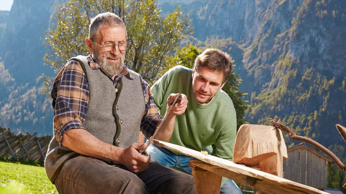 Ein älterer Mann schnitzt Holz beim Bauernherbst, während ein jüngerer Mann zusieht, mit den Bergen im Hintergrund.