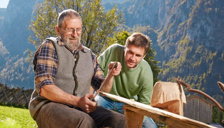 Ein älterer Mann schnitzt Holz beim Bauernherbst, während ein jüngerer Mann zusieht, mit den Bergen im Hintergrund.
