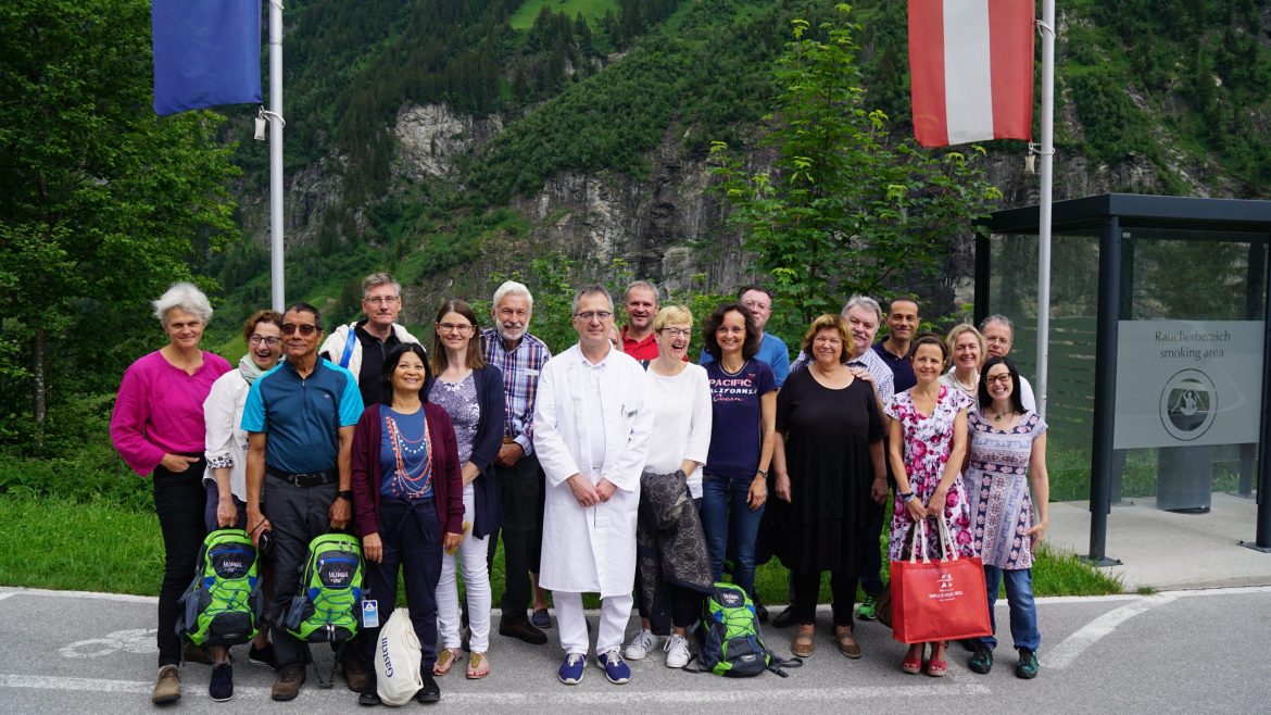 Eine Gruppe von Menschen auf einer Ärztestudienreise posiert im Freien in der Nähe von Fahnen und Bergen, im Hintergrund ist Grün zu sehen.