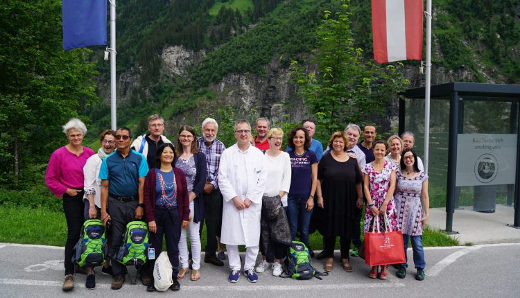 Eine Gruppe von Menschen auf einer Ärztestudienreise posiert im Freien in der Nähe von Fahnen und Bergen, im Hintergrund ist Grün zu sehen.