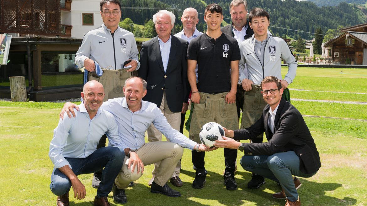 Eine Gruppe von Männern im Freien auf einer Wiese lächelt in die Kamera, zwei von ihnen halten einen Fußball in der Hand.