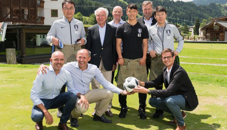Eine Gruppe von Männern im Freien auf einer Wiese lächelt in die Kamera, zwei von ihnen halten einen Fußball in der Hand.