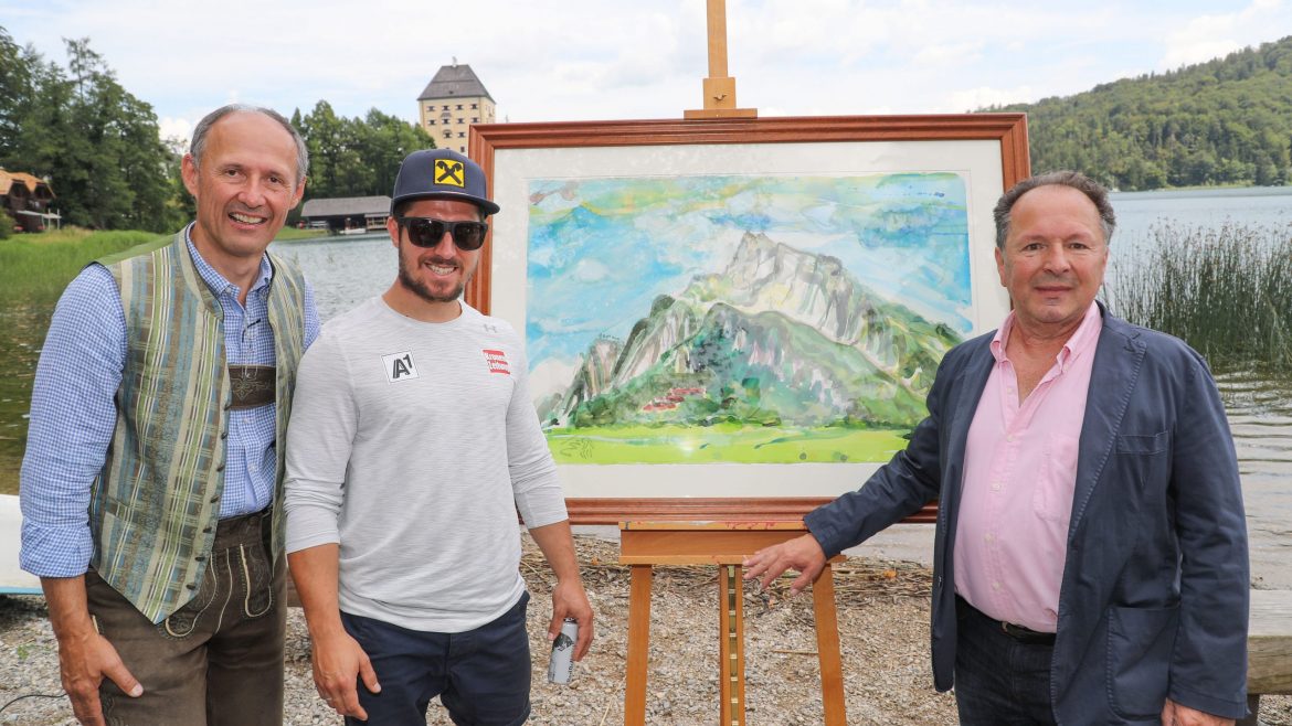 Drei Männer stehen an einem See, zwischen ihnen auf einer Staffelei ein buntes Berggemälde, inspiriert von Marcel Hirscher.