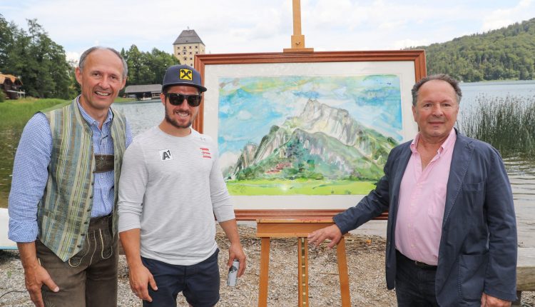 Drei Männer stehen an einem See, zwischen ihnen auf einer Staffelei ein buntes Berggemälde, inspiriert von Marcel Hirscher.