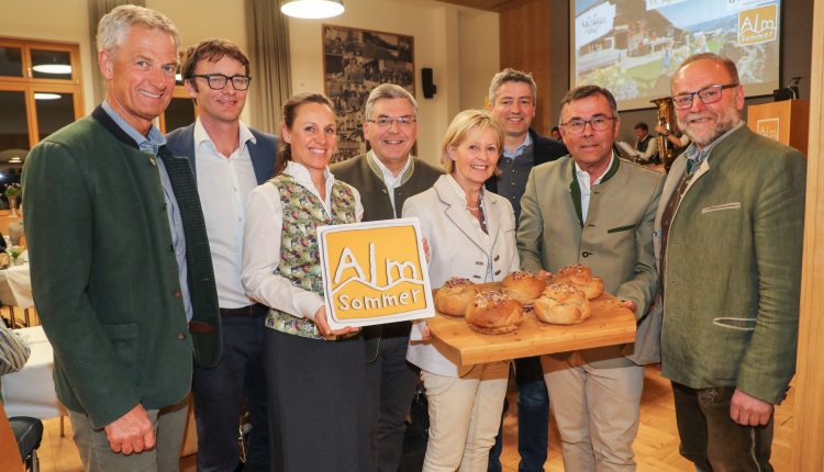 Acht Personen in österreichischer Tracht halten Brot und ein Almsommer-Schild bei einer Indoor-Veranstaltung.