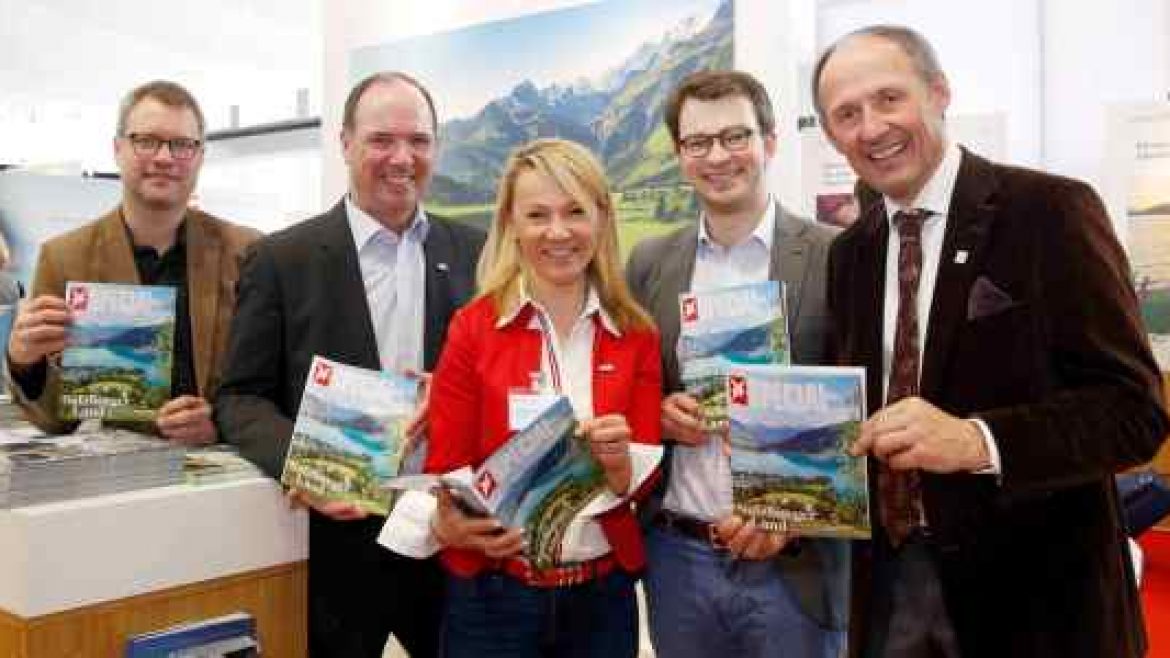 Fünf Menschen lächeln und halten Reisemagazine vor einer malerischen Schweizer Landschaft.