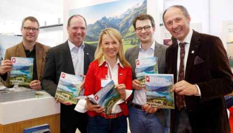 Fünf Menschen lächeln und halten Reisemagazine vor einer malerischen Schweizer Landschaft.