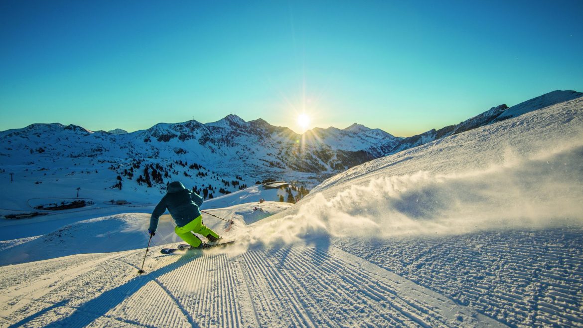 Ein Skifahrer gleitet bei Sonnenaufgang eine präparierte Schneepiste hinunter und genießt die Schönheit der winterlichen Bergkulisse.