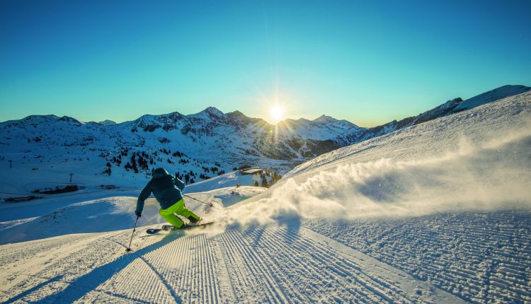 Ein Skifahrer gleitet bei Sonnenaufgang eine präparierte Schneepiste hinunter und genießt die Schönheit der winterlichen Bergkulisse.