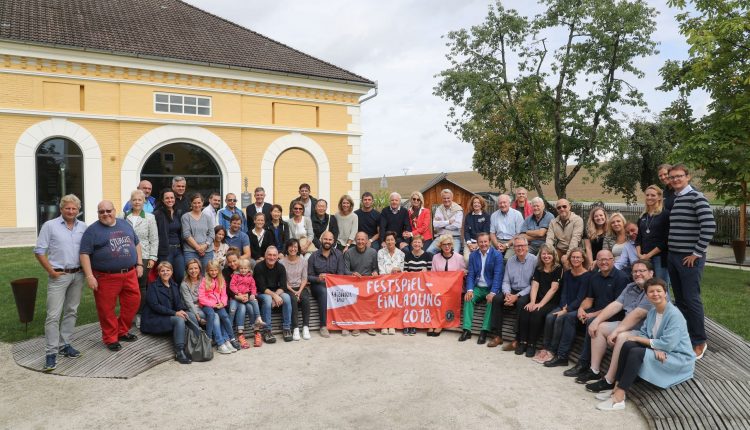 Eine große Gruppe lächelnder Menschen posiert im Freien und hält ein rotes Banner mit der Aufschrift "Festspiel Einladung 2018".