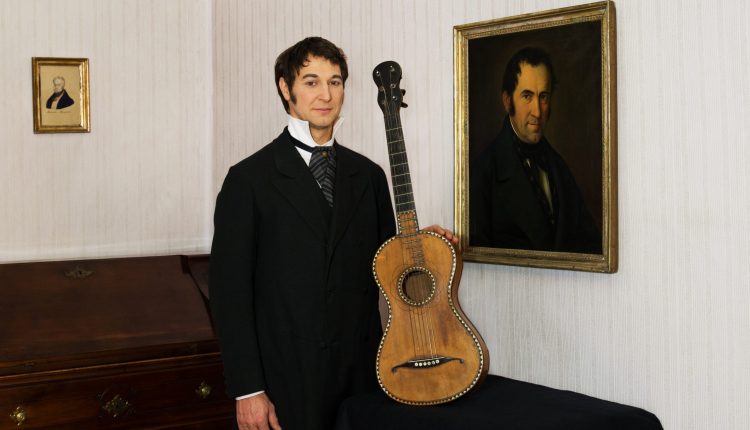 Ein Mann im Anzug steht neben einer antiken Gitarre und einem Porträt in einem Zimmer im Vintage-Stil.