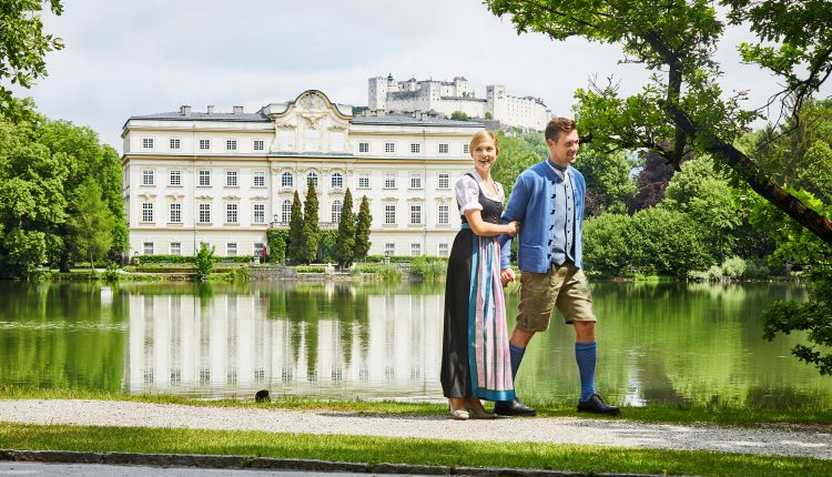 Ein Paar in traditioneller österreichischer Kleidung spaziert an einem See, mit einem großen Gebäude und einem Schloss im Hintergrund.