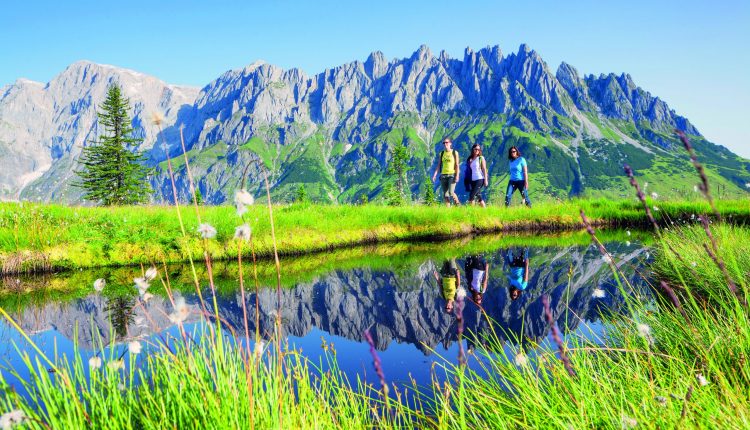 Vier Personen wandern an einem Teich, in dem sich die Berge und der blaue Himmel spiegeln.