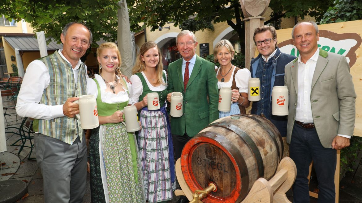 Sieben Personen in traditioneller österreichischer Kleidung halten Bierkrüge und stehen an einem hölzernen Bierfass im Freien.