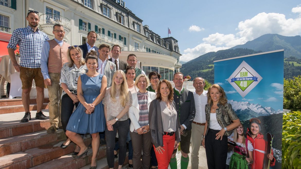 Eine Gruppe von Menschen posiert an einem sonnigen Tag gemeinsam vor einem Hotel mit Bergen im Hintergrund.