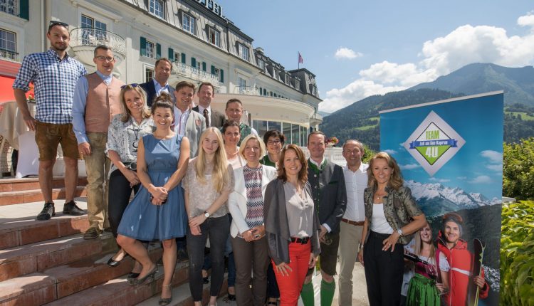Eine Gruppe von Menschen posiert an einem sonnigen Tag gemeinsam vor einem Hotel mit Bergen im Hintergrund.