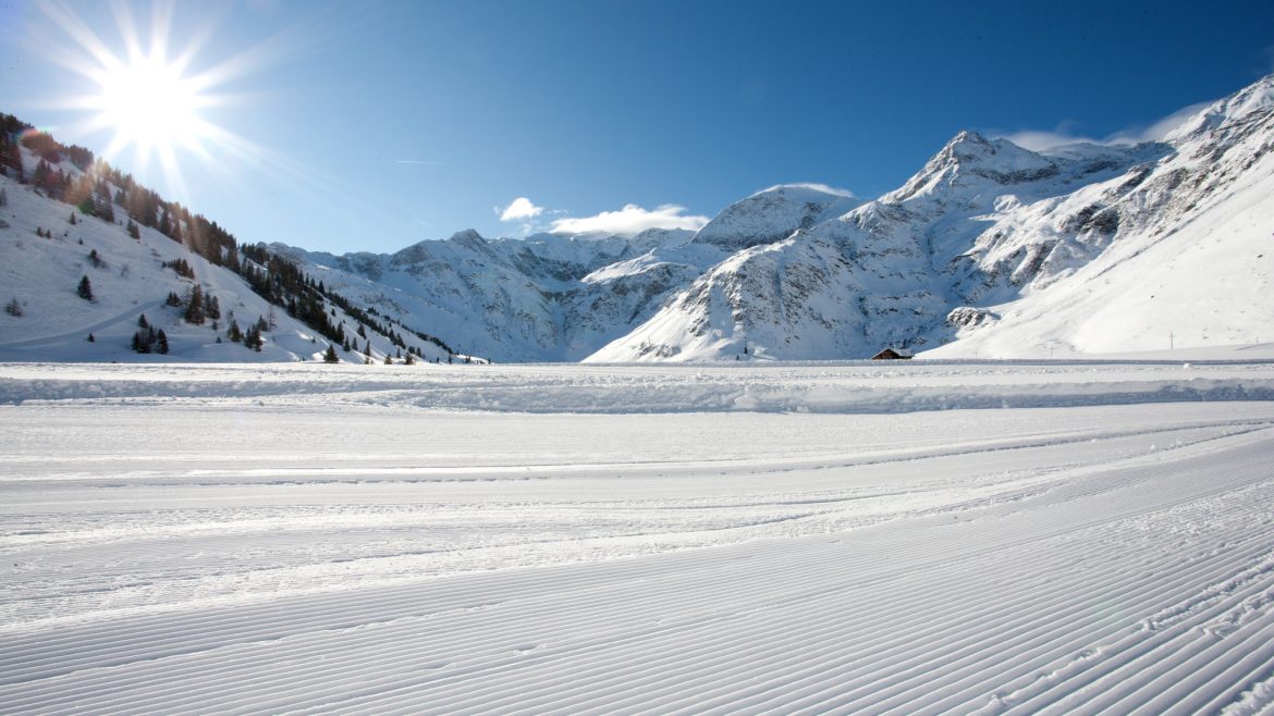 Die Sonne scheint auf eine verschneite Berglandschaft mit präparierten Skipisten im Vordergrund.