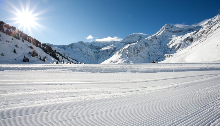 Die Sonne scheint auf eine verschneite Berglandschaft mit präparierten Skipisten im Vordergrund.