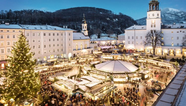 Ein festlicher Weihnachtsmarkt in der Abenddämmerung mit Lichtern, einem großen Baum und vielen Menschen auf einem verschneiten Stadtplatz.