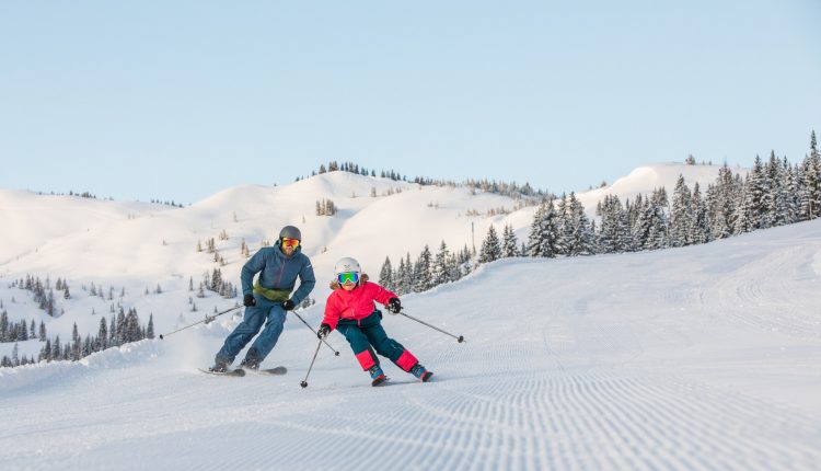Ein Erwachsener und ein Kind fahren auf einer präparierten, verschneiten Piste mit Kiefern und Bergen im Hintergrund.
