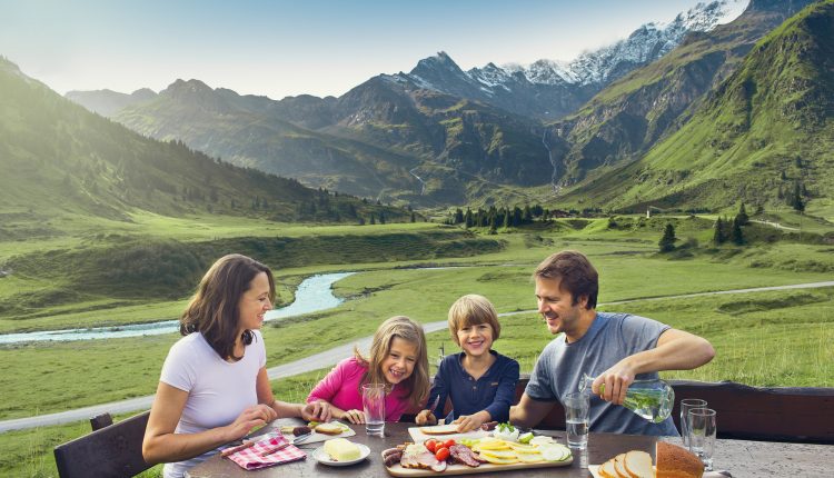 Eine Familie genießt ein Picknick an einem Holztisch in einem malerischen Bergtal mit einem Fluss und schneebedeckten Gipfeln.