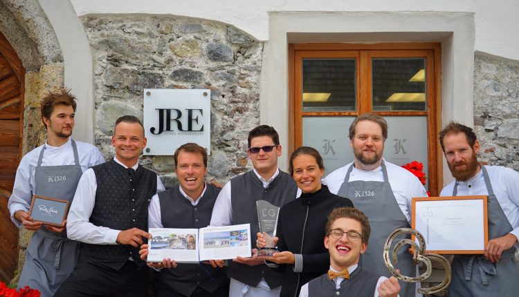 Ein lächelndes Restaurantteam posiert mit Auszeichnungen und Urkunden vor einem Steingebäude mit einem JRE-Schild.