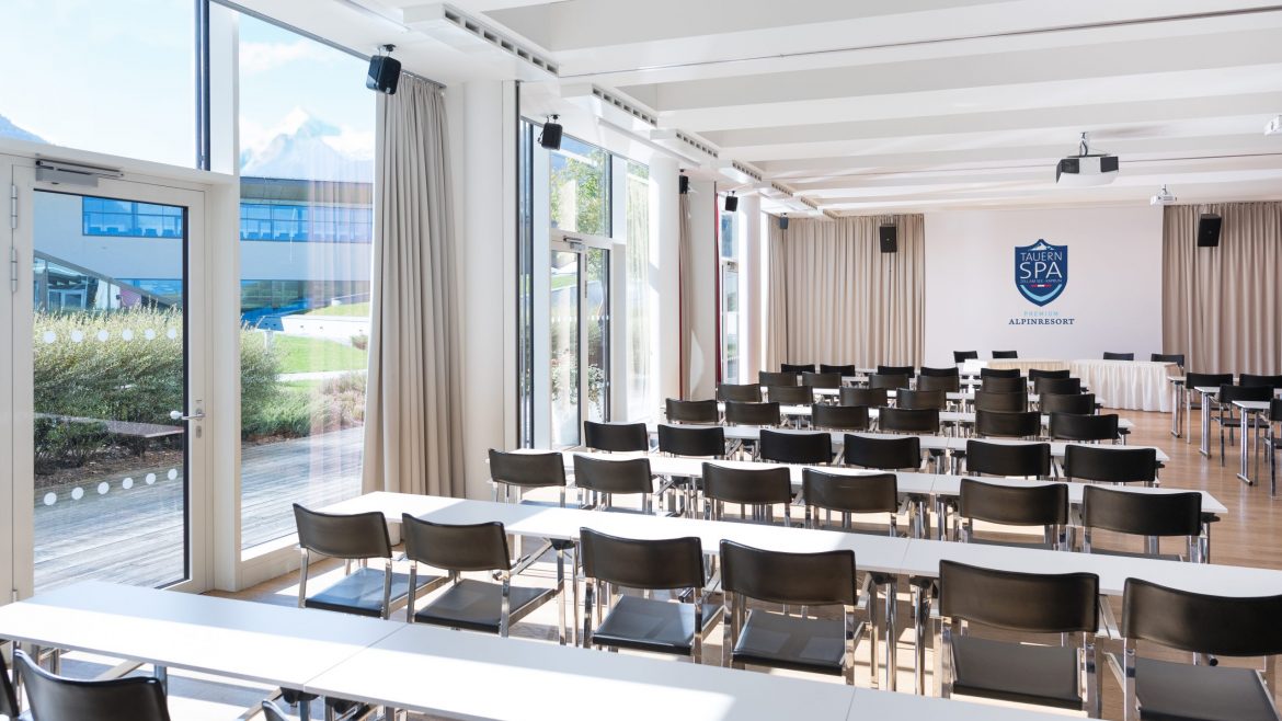 Heller Konferenzraum mit Stuhlreihen, Tischen, großen Fenstern und einem SPA ACADEMY-Schild an der Stirnseite.