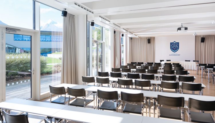Heller Konferenzraum mit Stuhlreihen, Tischen, großen Fenstern und einem SPA ACADEMY-Schild an der Stirnseite.