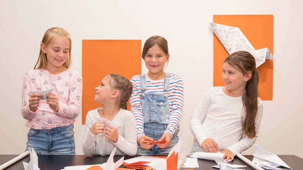 Vier junge Mädchen lächeln, während sie an einem Tisch mit orangefarbenem Papier und Origami-Kunstwerken an der Wand hinter ihnen Papier-Origami basteln.