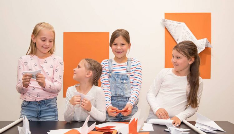 Vier junge Mädchen lächeln, während sie an einem Tisch mit orangefarbenem Papier und Origami-Kunstwerken an der Wand hinter ihnen Papier-Origami basteln.
