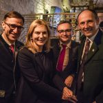 Vier Personen in formeller Kleidung, die bei einer Veranstaltung in einem Innenraum mit warmer Beleuchtung lächelnd zusammen posieren.