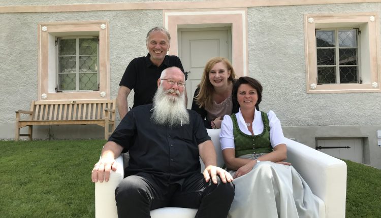 Vier Erwachsene posieren und lächeln im Freien auf einer weißen Couch vor einem hellen Gebäude mit Fenstern.