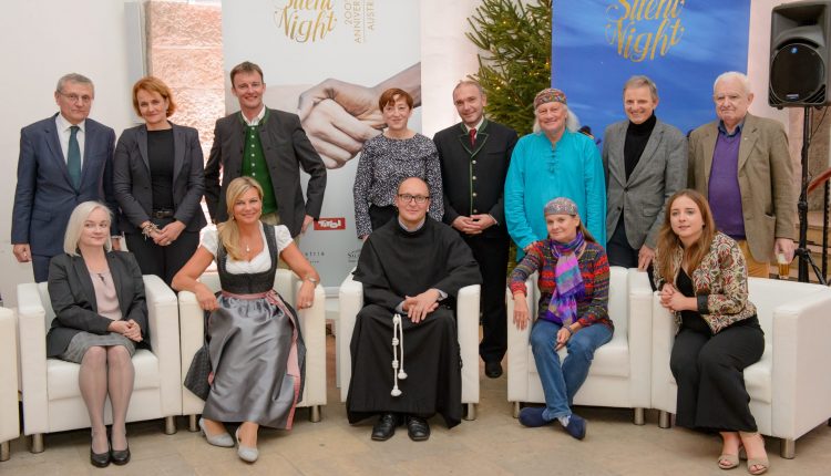 Dreizehn Personen posieren in einem Haus mit Stille-Nacht-Bannern und einem kleinen Baum im Hintergrund.