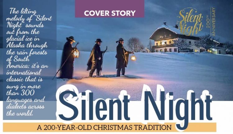 Drei Menschen mit Laternen gehen im Schnee auf ein beleuchtetes Haus zu, das nachts für die Titelgeschichte der Weihnachtstradition Stille Nacht erleuchtet ist.