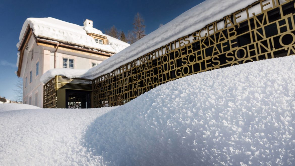 Ein schneebedecktes Gebäude mit einem modernen goldenen Metallzaun mit ausgeschnittenem Text unter einem klaren blauen Himmel.