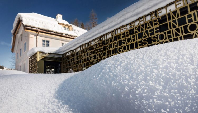 Ein schneebedecktes Gebäude mit einem modernen goldenen Metallzaun mit ausgeschnittenem Text unter einem klaren blauen Himmel.