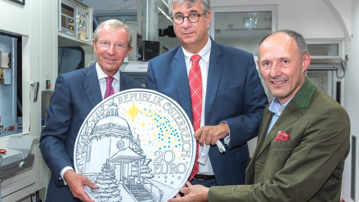 Drei Männer in Anzügen halten eine große österreichische 20-Euro-Gedenkmünze in einer hellen, industriellen Umgebung.