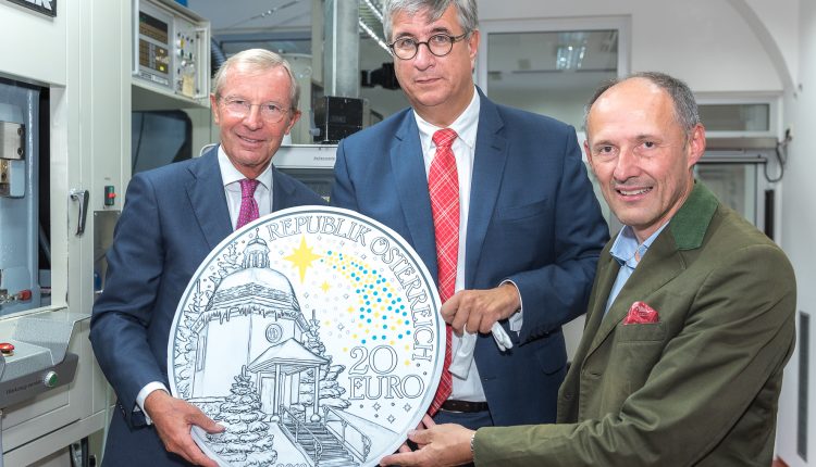 Drei Männer in Anzügen halten eine große österreichische 20-Euro-Gedenkmünze in einer hellen, industriellen Umgebung.
