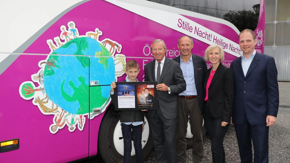 Fünf Personen stehen an einem rosa-weißen Bus; ein Junge hält eine Urkunde und ein Plakat mit vier Erwachsenen.
