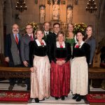 Neun Erwachsene in traditioneller formeller Kleidung posieren in einer geschmückten Kirche mit verziertem Altar und Statuen.