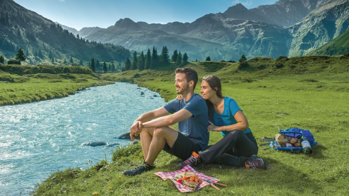 Ein Paar sitzt an einem Fluss in einem Bergtal und genießt ein Picknick im grünen Gras unter einem klaren Himmel.