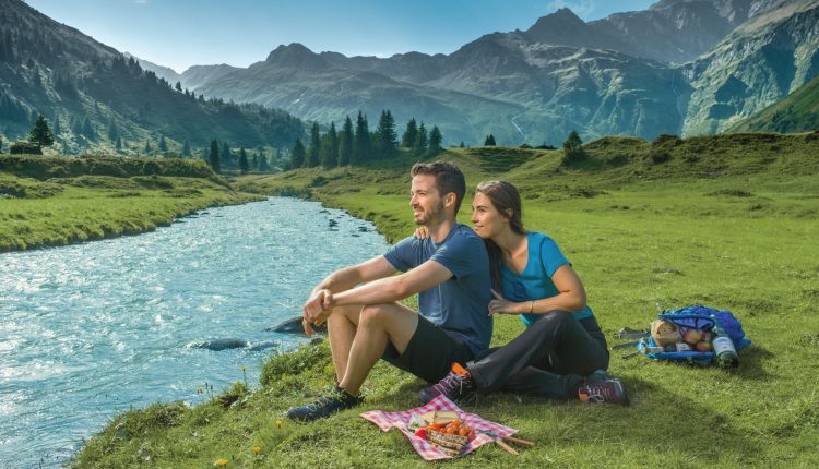 Ein Paar sitzt an einem Fluss in einem Bergtal und genießt ein Picknick im grünen Gras unter einem klaren Himmel.